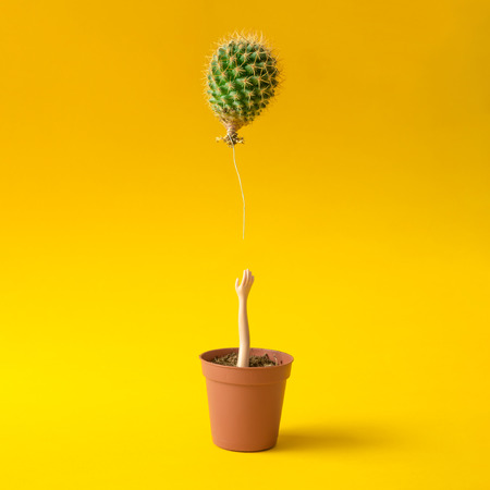 Doll hand reaching for cactus balloon out of flower pot on yellow background. Creative minimal concept.の写真素材