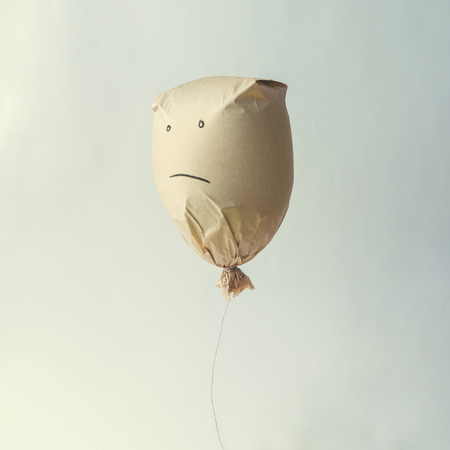 Paper bag balloon with sad face emoji on white background. Creative minimal concept.の写真素材