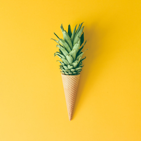 Ice cream cone with pineapple leaves on bright yellow background. Fruit and candy concept. Flat lay.