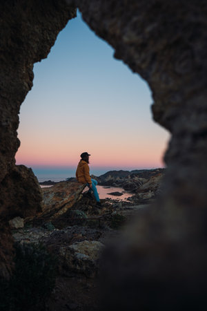 A girl sitting on a rock with a sunset at the blue hour between a rock in Cap de Creus, Spainの写真素材