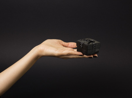 Female hand holding a black gift box decorated with black ribbon with white polka dots on it. Isolated on black background.の写真素材