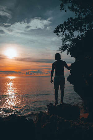 The guy enjoys the sunset riding on a swing on the ptropical beach. Silhouettes of a guy on a swing hanging on a palm treeの写真素材