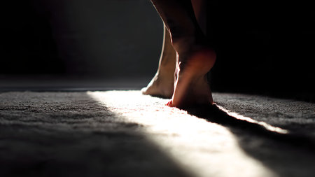 Bare feet walking on a carpet in a dark room with a single light source. Conceptual image of loneliness.の素材