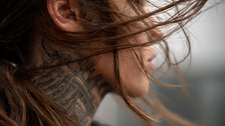 Close-up side view of a woman with long brown hair blowing in the wind, showcasing a detailed neck tattoo.の素材