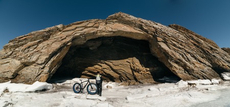 Fatbike also called fat bike or fat-tire bike - Cycling on large wheels. Traveller cyclist standing on the ice of Lake Baikal, on the background of the island with a small cave gorge .の写真素材