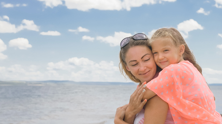 Little cute girl baby and young mother at beach. Mom sits on the shore, and her daughter is coming from behind. The kid hugs and kisses her mother. Mother and her child.の写真素材