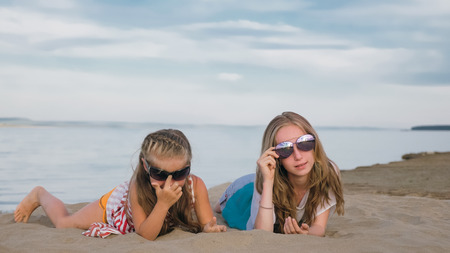 Two teenage are sitting on a sandy beach near the sea. Girl playing, talk to each other. Sisters are dressed in dresses and sunglasses. Children have real emotions of happiness.の写真素材