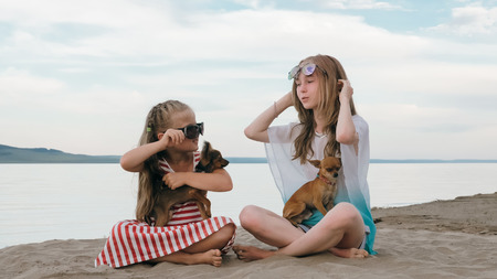 Two teenage are sitting on a sandy beach near the sea. Girl playing, talk to each other. They have two dogs. Children have real emotions of happiness.の写真素材