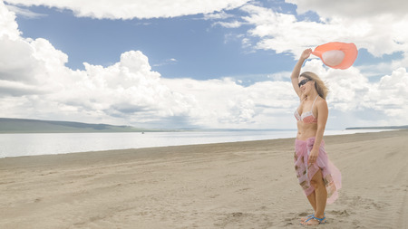 Attractive woman in a hat stands on the beach against the blue sea. She has sunglasses and a  on her head. On her body is a swimsuit and a silk cape. In the background are very beautiful deep clouds.の写真素材