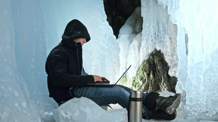 Hacker hacks the server. Man to program on laptop in ice cave. Around the mysterious ice grotto. Man hides himself in a hood. Hacker has criminal cases on the Internet. Guy is warming himself with the coffee from the thermos. Hes a cool hipster.の写真素材