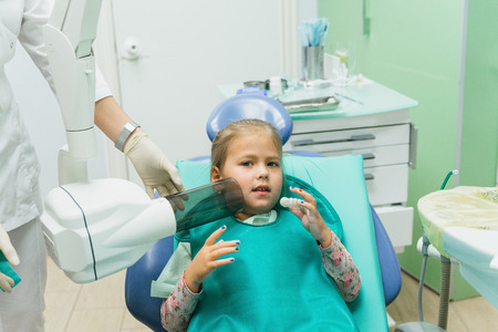 Child with a mother at a dentists reception. The girl lies in the chair, behind her mother. The doctor works with an assistant. Procedure for drilling a tooth. Setting a temporary seal. Using the x-ray machine.の写真素材