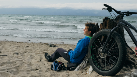 Fat bike also called fatbike or fat-tire bike in summer driving on the beach. The guy sits on the wheel of his bicycle, looks at the sea.の写真素材