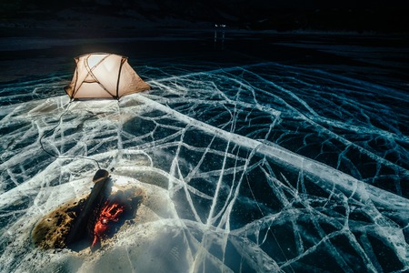 Fire on ice at night. Campground on ice. Tent stands next to bonfire. Lake Baikal. Nearby there is car. Shelter tent and ice are illuminated from the inside. Beautiful bonfire on real cracked ice. Moonlight night.の写真素材