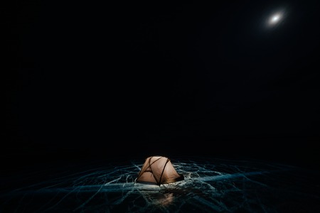 Fire on ice at night. Campground on ice. Tent stands next to bonfire. Lake Baikal. Nearby there is car. Shelter tent and ice are illuminated from the inside. Beautiful bonfire on real cracked ice. Moonlight night.の写真素材