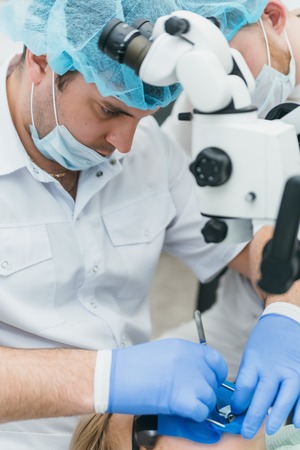 Doctor used microscope. Dentist is treating patient in modern dental office. Operation is carried out using cofferdam. Client is inserted and restored teeth, make denture. Orthodontist and assistant work in protective masks.の写真素材