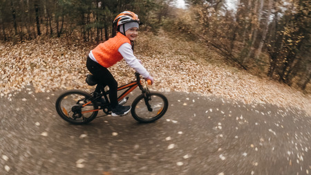 One caucasian children rides bike road in autumn park. Little girl riding black orange cycle in forest. Kid goes do bicycle sports. Biker motion ride with backpack and helmet. Mountain bike hardtail.の写真素材