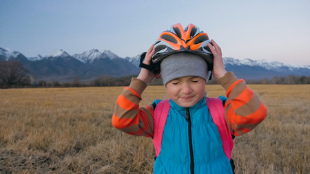 One caucasian children walk with bike in wheat field. Little girl walking black orange cycle on background of beautiful snowy mountains. Biker stand with backpack and helmet. Mountain bike hardtail.の写真素材