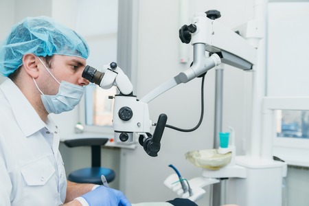 Doctor used microscope. Dentist is treating patient in modern dental office. Operation is carried out using cofferdam. Client is inserted and restored teeth, make denture. Orthodontist and assistant work in protective masks.の写真素材