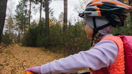 One caucasian children walk with bike in autumn park. Little girl walking black orange cycle in forest. Kid goes do bicycle sports. Biker motion ride with backpack and helmet. Mountain bike hardtail.の写真素材
