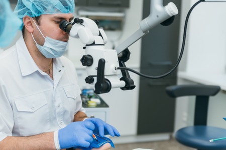 Doctor used microscope. Dentist is treating patient in modern dental office. Operation is carried out using cofferdam. Client is inserted and restored teeth, make denture. Orthodontist and assistant work in protective masks.の写真素材