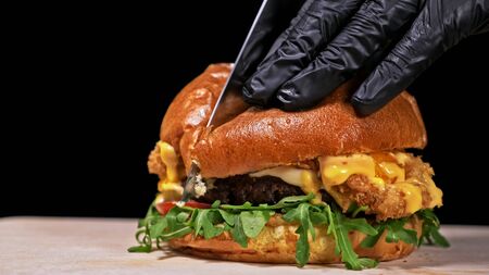 Cut craft burger is cooking on black background in black food gloves. Consist: sauce, arugula,...