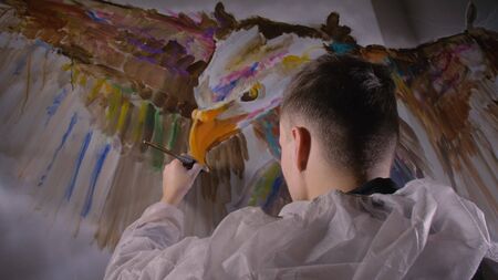 Artist designer draws an eagle on the wall. Craftsman decorator paints a picture with acrylic oil color. Painter painter dressed in a paint coat. Indoor. Dark magic cinematic look.の写真素材