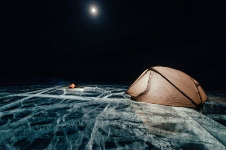 Fire on ice at night. Campground on ice. Tent stands next to bonfire. Lake Baikal. Nearby there is car. Shelter tent and ice are illuminated from the inside. Beautiful bonfire on real cracked ice. Moonlight night.の写真素材