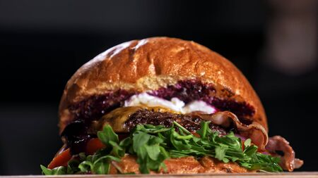 Craft burger is cooking on black background. Consist: sauce, arugula, tomato, red onion, bacon, red currant sauce, ricotta cottage fresh cheese, air bun and marble meat beef. Not made ideal. Looks real, loving hand made.の写真素材