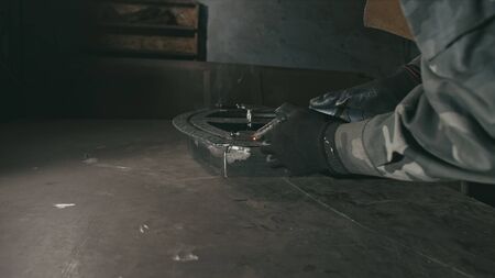 Forge workshop. Smithy. Worker in a welding hood helmet welds a part by electric welding. Sparks are...