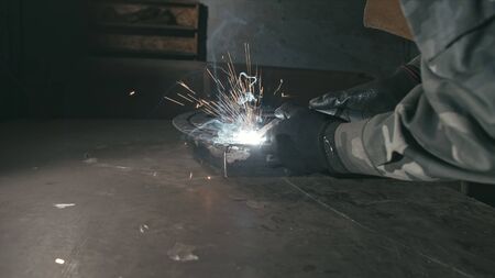 Forge workshop. Smithy. Worker in a welding hood helmet welds a part by electric welding. Sparks are...