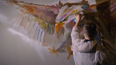 Artist designer draws an eagle on the wall. Craftsman decorator paints a picture with acrylic oil color. Painter painter dressed in a paint coat. Indoor. Dark magic cinematic look.の写真素材