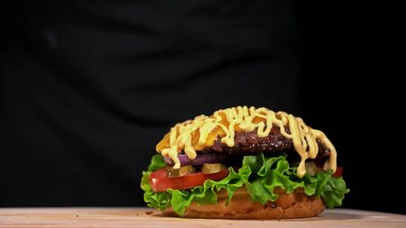 Craft burger is cooking on black background in black food gloves. Consist: sauce, lettuce, tomato, red onion, pickle, cheese, bacon, air bun and marbled meat beef. Not made ideal. Looks real, loving hand made.の写真素材