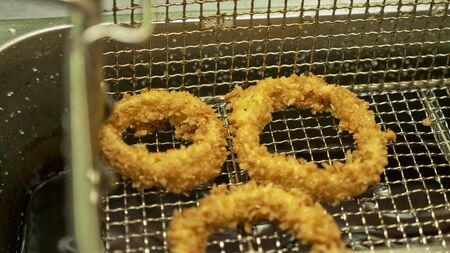 Deep fried onion ring. Oniony Rings cooking in hot bubbly oil in a deeped fryer. Golden chopped onions circle frying process in a wire metal basket hot oils in a restaurant kitchen viewed closeup from above.の写真素材