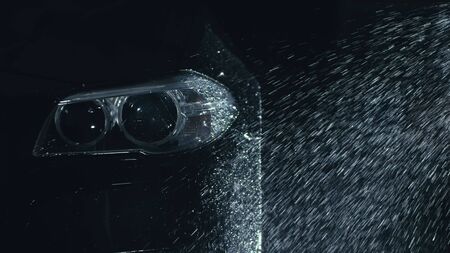 Car headlight wash. Washing modern vehicle body by high pressure jet wash hose water. Auto glass headlamp, angel eyes in drops. Close up of the spotlight of a black automobile as it gets cleaned.の写真素材