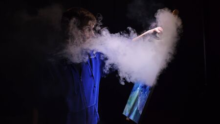 Artist copyist paint seascape with ship in ocean. Vaper smoke vape e-cigarette. Craftsman decorator draw as boat sail on blue sea with acrylic oil color. Draw finger, brush, knife palette. Indoor.の写真素材