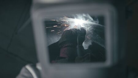 Forge workshop. Smithy. Worker in a welding hood helmet welds a part by electric welding. Sparks are reflected in the protective screen. Blacksmith makes iron product for manufacture. Slow motion.の写真素材
