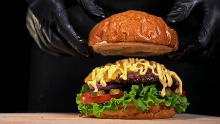 Craft burger is cooking on black background in black food gloves. Consist: sauce, lettuce, tomato, red onion, pickle, cheese, bacon, air bun and marbled meat beef. Not made ideal. Looks real, loving hand made.の写真素材