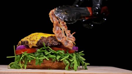 Craft burger is cooking on black background. Consist: sauce, arugula, tomato, red onion, bacon, red currant sauce, ricotta cottage fresh cheese, air bun and marble meat beef. Not made ideal. Looks real, loving hand made.の写真素材