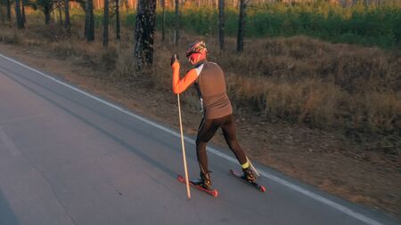 Training an athlete on the roller skaters. Biathlon ride on the roller skis with ski poles, in the helmet. Autumn workout. Roller sport. Adult man riding on skates.の写真素材
