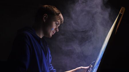 Artist copyist paint seascape with ship in ocean. Vaper smoke vape e-cigarette. Craftsman decorator draw as boat sail on blue sea with acrylic oil color. Draw finger, brush, knife palette. Indoor.の写真素材