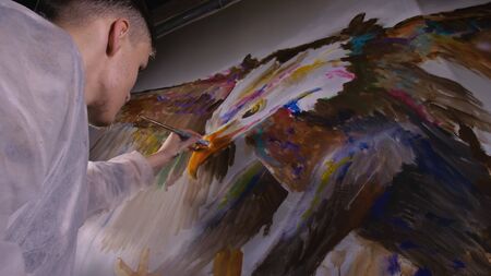 Artist designer draws an eagle on the wall. Craftsman decorator paints a picture with acrylic oil color. Painter painter dressed in a paint coat. Indoor. Dark magic cinematic look.の写真素材