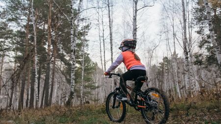 One caucasian children rides bike road in autumn park. Little girl riding black orange cycle in forest. Kid goes do bicycle sports. Biker motion ride with backpack and helmet. Mountain bike hardtail.の写真素材