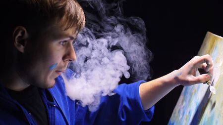 Artist copyist paint seascape with ship in ocean. Vaper smoke vape e-cigarette. Craftsman decorator draw as boat sail on blue sea with acrylic oil color. Draw finger, brush, knife palette. Indoor.の写真素材