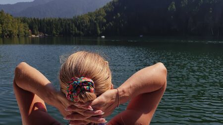 Woman sit on a sunbed in sunglasses and swimming suit. Girl rest on a flood wood underwater pier. The pavement is covered with water in lake. Background are mountain and forest. Swimwear bikini blue.の写真素材
