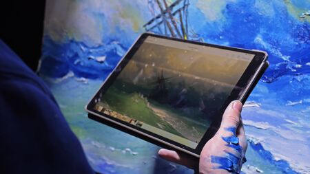 Artist copyist paint seascape with ship in ocean. Craftsman decorator draw as boat sail on blue sea with acrylic oil color. Draw finger, brush, knife palette. Indoor. In hands of tablet computer pc.の写真素材