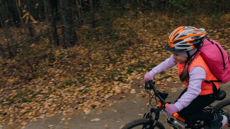 One caucasian children rides bike road in autumn park. Little girl riding black orange cycle in forest. Kid goes do bicycle sports. Biker motion ride with backpack and helmet. Mountain bike hardtail.の写真素材