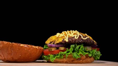 Craft burger is cooking on black background in black food gloves. Consist: sauce, lettuce, tomato, red onion, pickle, cheese, bacon, air bun and marbled meat beef. Not made ideal. Looks real, loving hand made.の写真素材