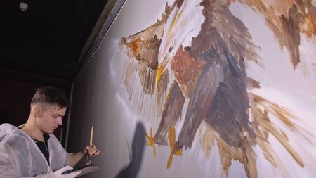 Artist designer draws an eagle on wall. Craftsman decorator paints picture with acrylic oil color looking at sketch in phone. Painter painter dressed in paint coat. Indoor. Dark magic cinematic look.の写真素材
