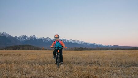 One caucasian children rides bike in wheat field. Little girl riding black orange cycle on background of beautiful snowy mountains. Biker motion ride with backpack and helmet. Mountain bike hardtail.の写真素材