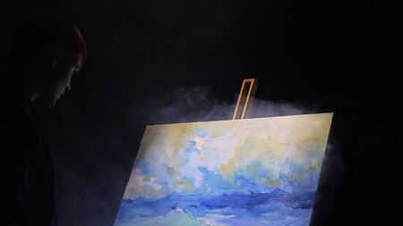 Artist copyist paint seascape with ship in ocean. Vaper smoke vape e-cigarette. Craftsman decorator draw as boat sail on blue sea with acrylic oil color. Draw finger, brush, knife palette. Indoor.の写真素材